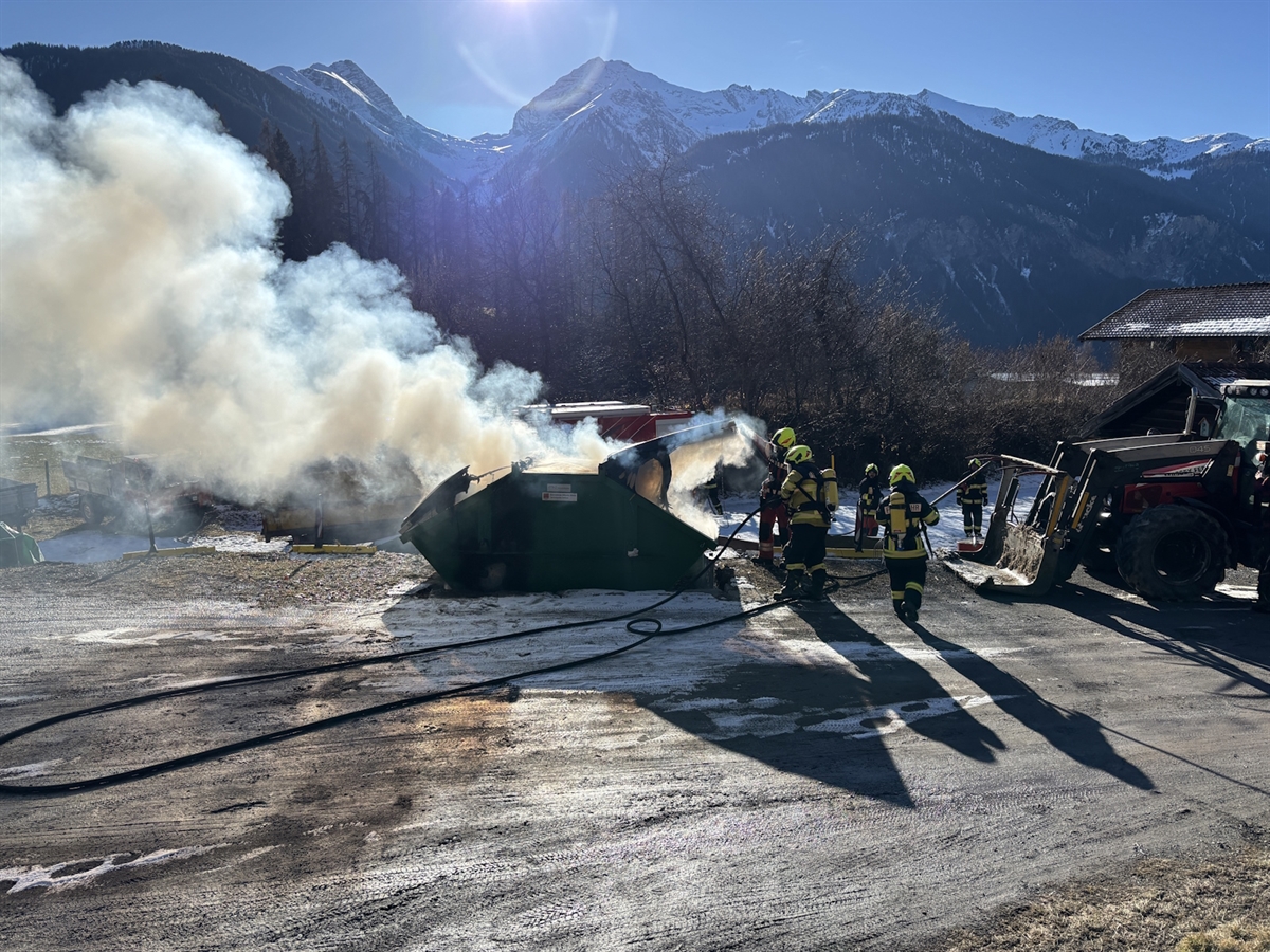 Aus der Mulde steigt Rauch empor. Feuerwehrleute beim Löscheinsatz. In der Ferne Bergpanorama.
