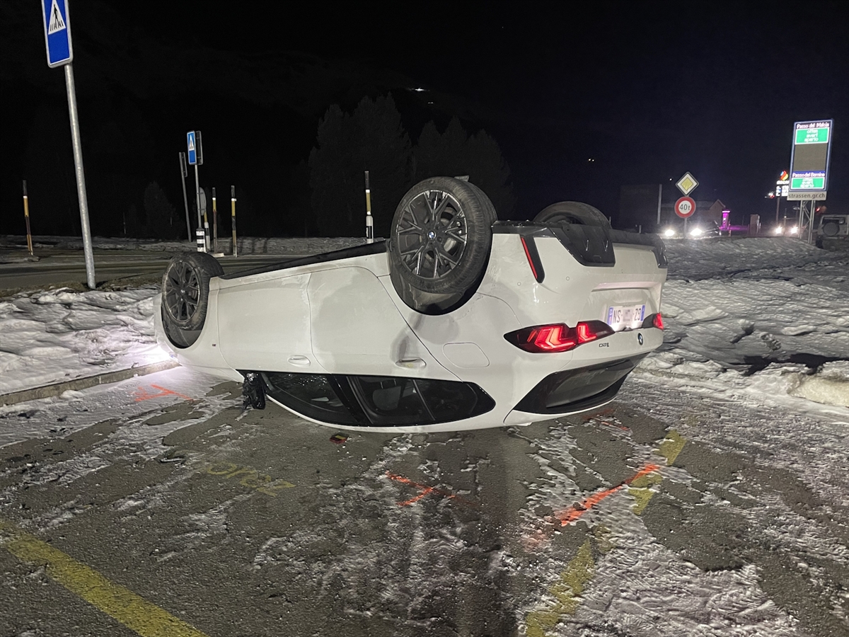 Der weisse BMW liegt auf dem Dach auf dem Parkplatz neben dem Kreisel. Im Hintergrund reflektiert im Scheinwerferlicht eine Informationstafel.
