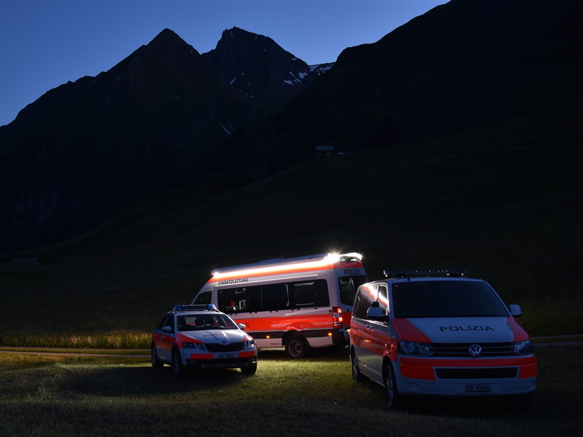 Aufnahme in der Dämmerung. Drei Einsatzfahrzeuge der Polizei stehen auf einer Wiese. Eines beleuchtet die nahe Umgebung. Im Hintergrund heben sich Berge vom wolkenlosen Himmel ab.