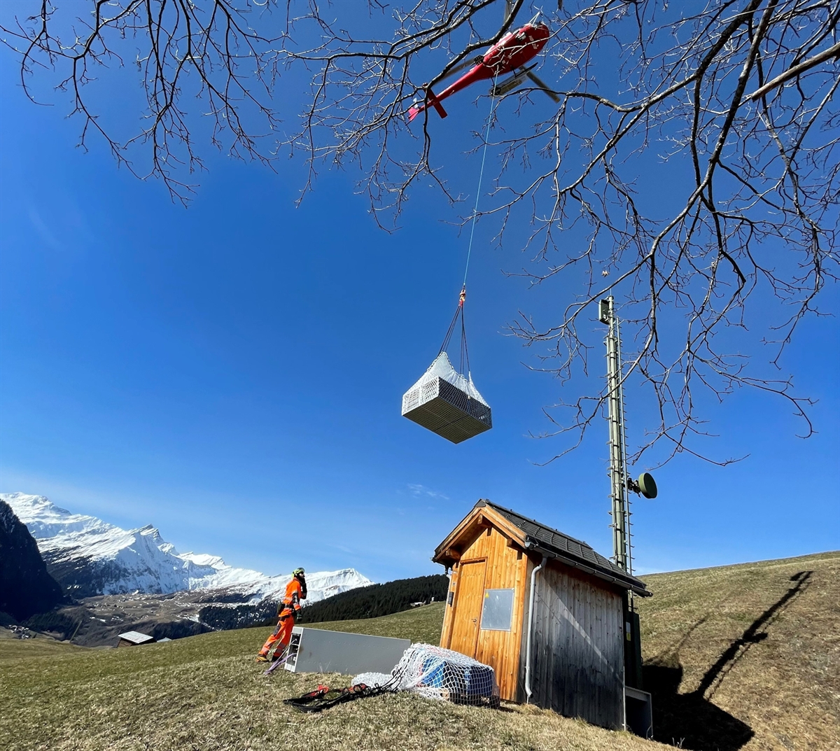 Ein Helikopter fliegt Material zu der Basis und Richtfunkstation Lumbrein. Am Boden ein Flughelfer, der den Abladeort dem Pilot weitergibt. Das Bild wird mit verschneiten Bergen sowie blauem Himmel abgerundet.